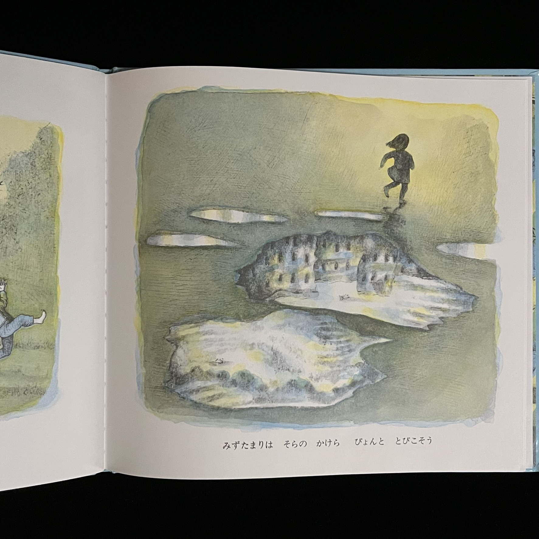 本の中のページの写真。子どもが遊ぶかたわらの水たまりができ、そこに空や雲、建物が映りこんでいる