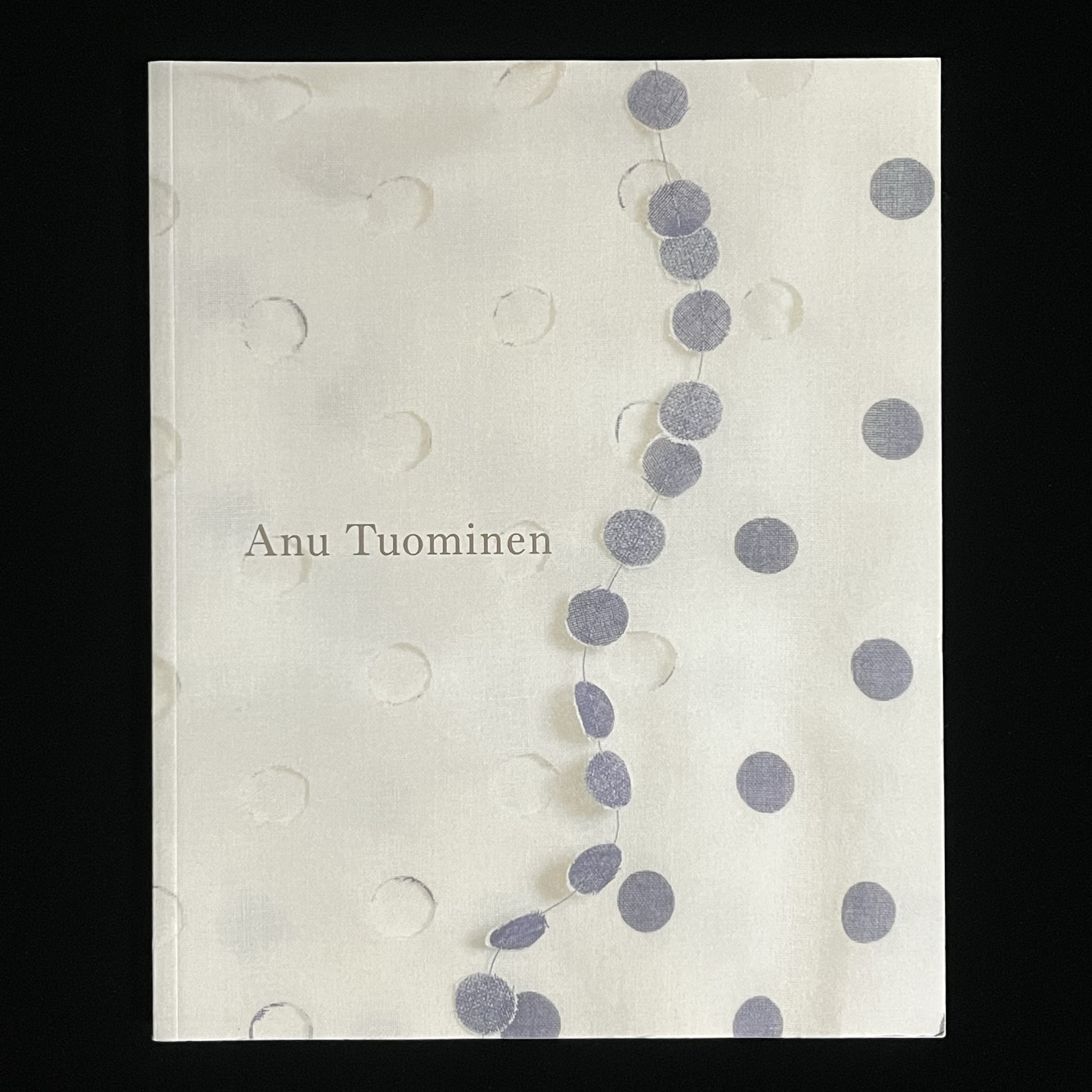『Anu Tuominen』の本の表紙の写真。切り抜かれたグレー丸い布が糸でつながれていて、切り抜かれた布がその下に敷かれています。この写真はお洋服の一部を拡大したもので、下に敷かれているのはお洋服の生地、切り抜かれた丸のつながりはネックレスになっています。